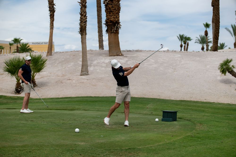 Two golfers on a desert golf course with palm trees, one mid-swing with a driver on a green fairway.