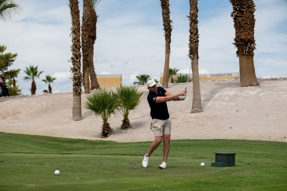 Golfer swinging a club on a desert golf course with palm trees and sandy terrain under a cloudy sky.