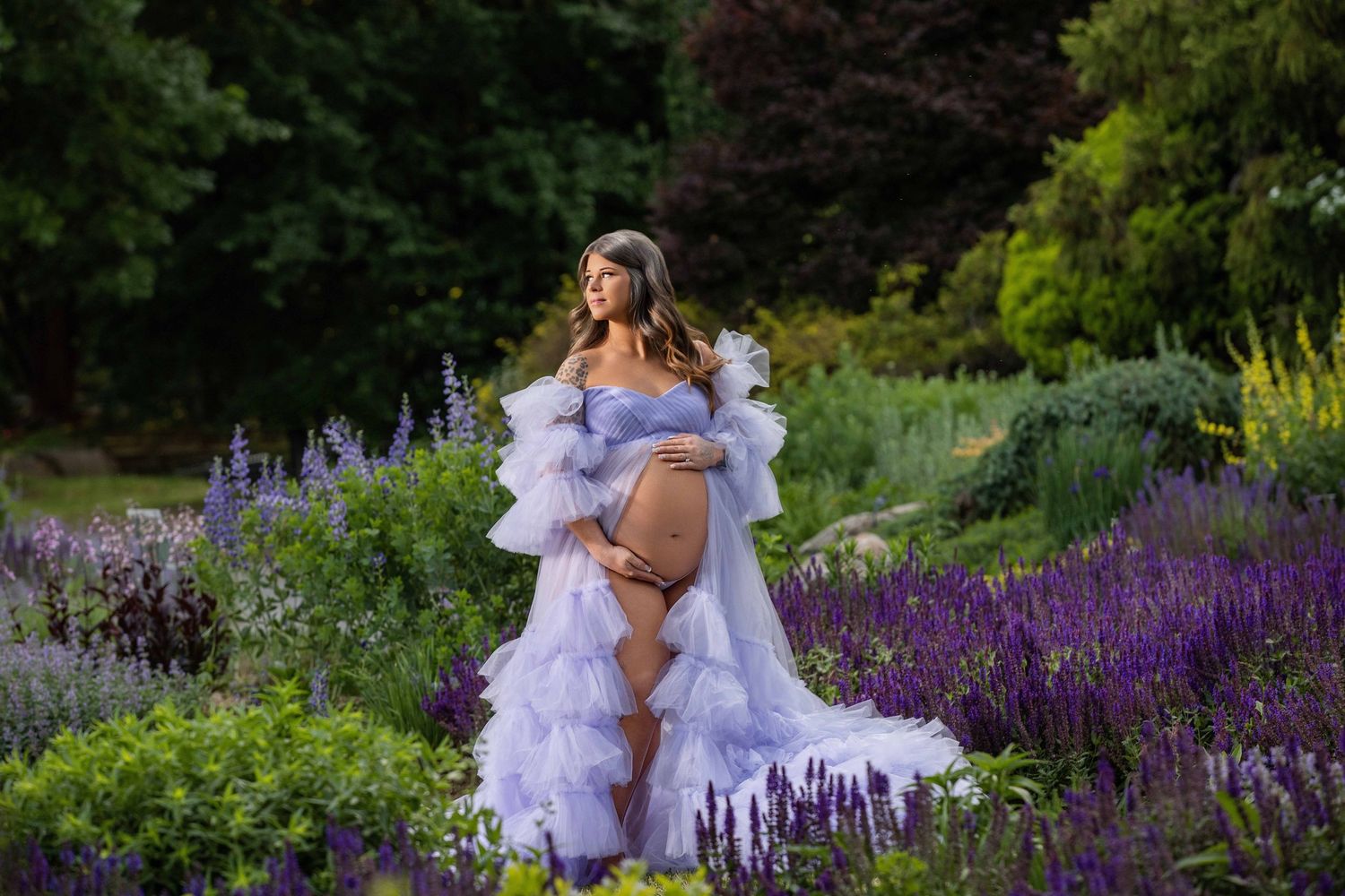 Pregnant woman in lavender tulle maternity gown poses among purple lavender flowers in lush garden setting.