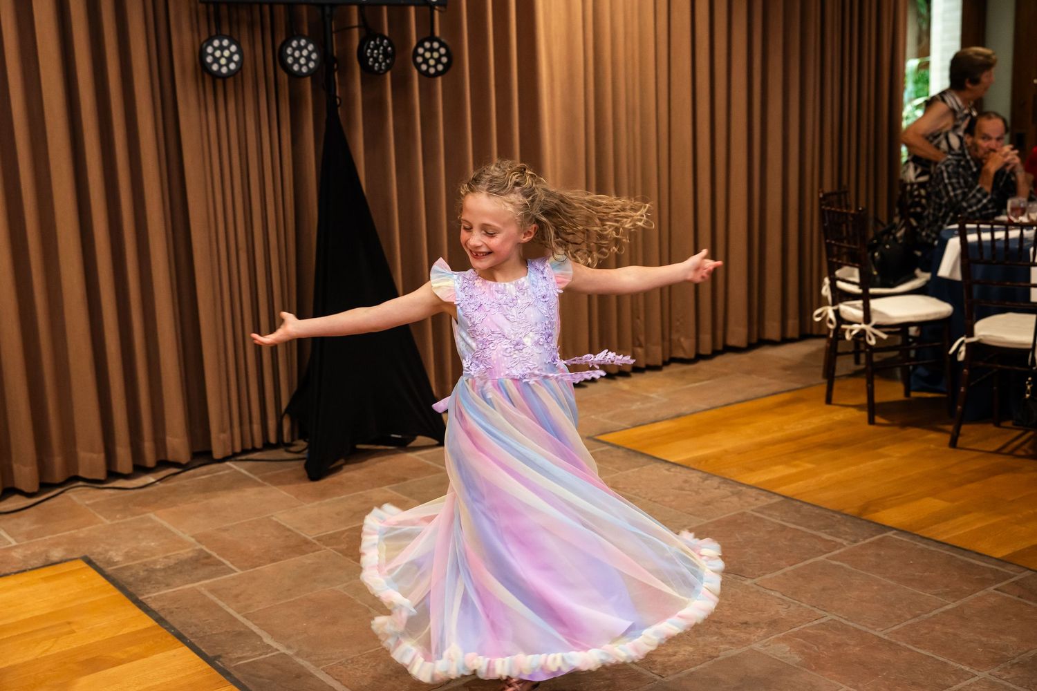 A young girl in a colorful pastel rainbow dress twirls joyfully on a dance floor at an indoor event.