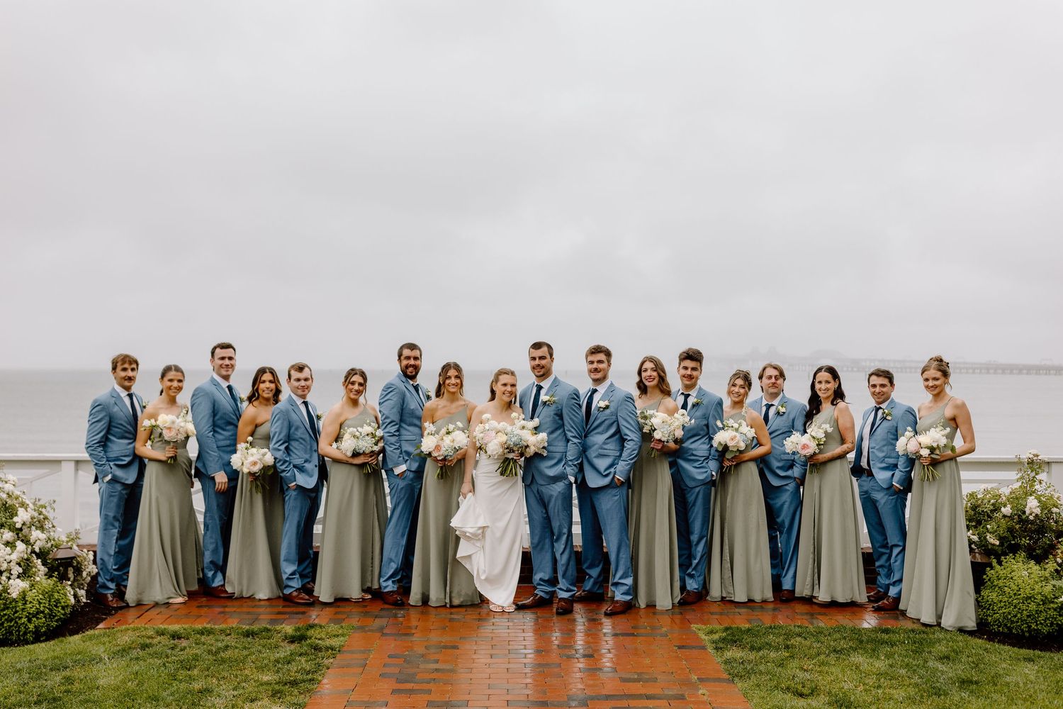 Wedding party in blue suits and sage green gowns posing outdoors by the ocean on a cloudy day.