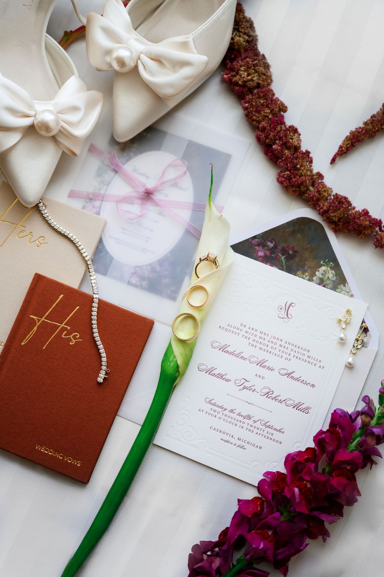 Elegant wedding flat lay with ivory bow heels, floral invitations, gold rings, pearl bracelet, and burgundy flowers on white fabric.