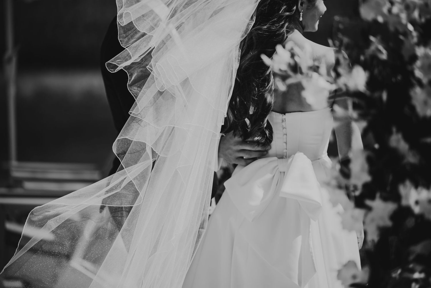 Black and white wedding photo of bride in elegant gown with flowing veil, embracing groom outdoors near lush greenery.