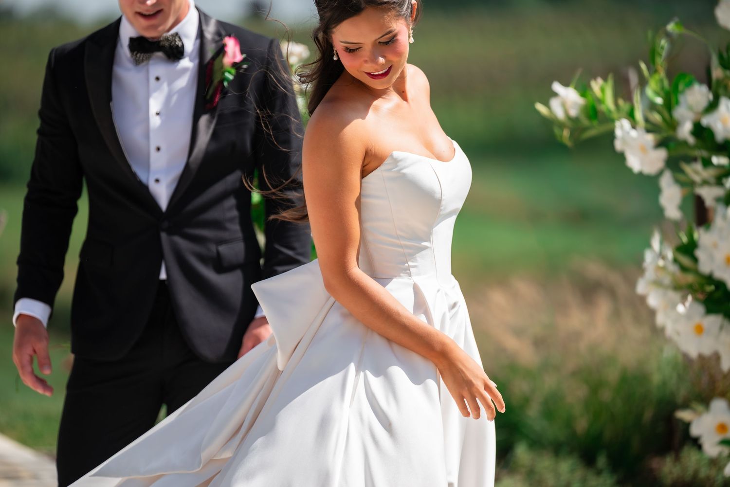 Bride in strapless white gown twirls beside groom in black tuxedo during outdoor wedding ceremony.