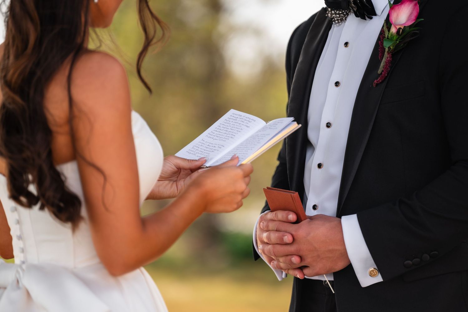 Bride reading wedding vows from paper to groom in tuxedo during outdoor ceremony.