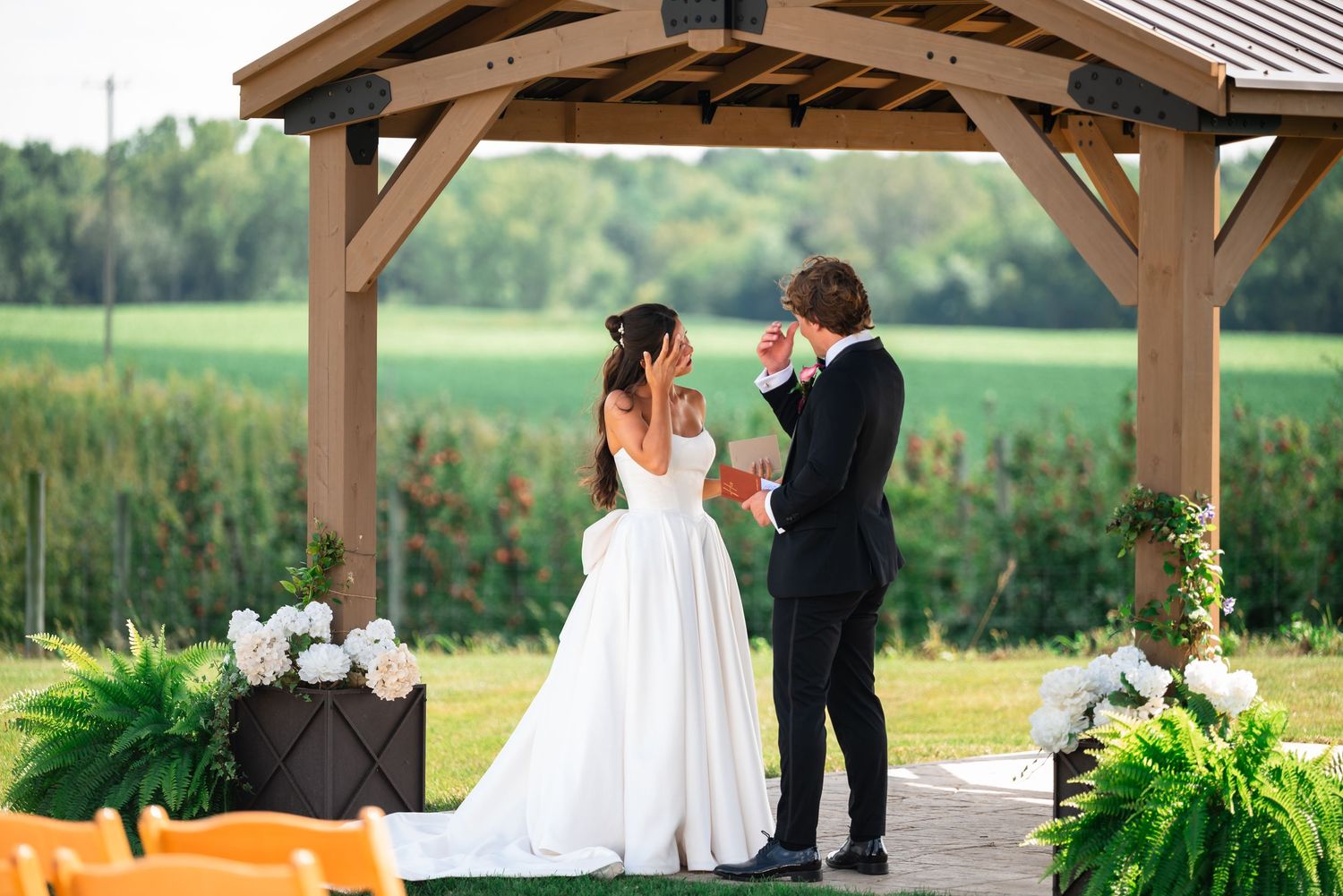Bride and groom exchanging vows under a wooden gazebo surrounded by lush green fields at an outdoor wedding ceremony.