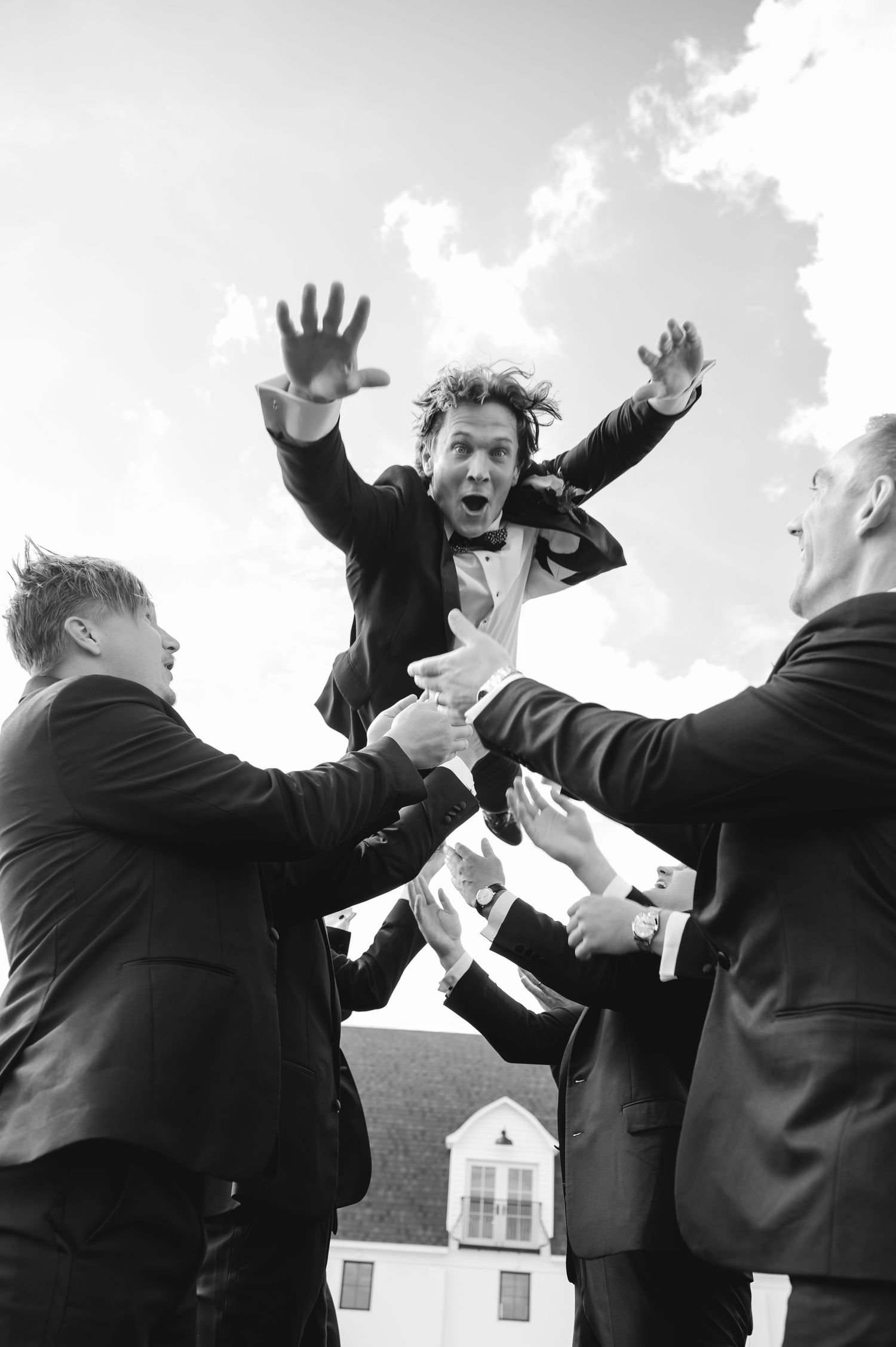 Groomsmen toss groom into air in black and white wedding celebration photo outdoors.