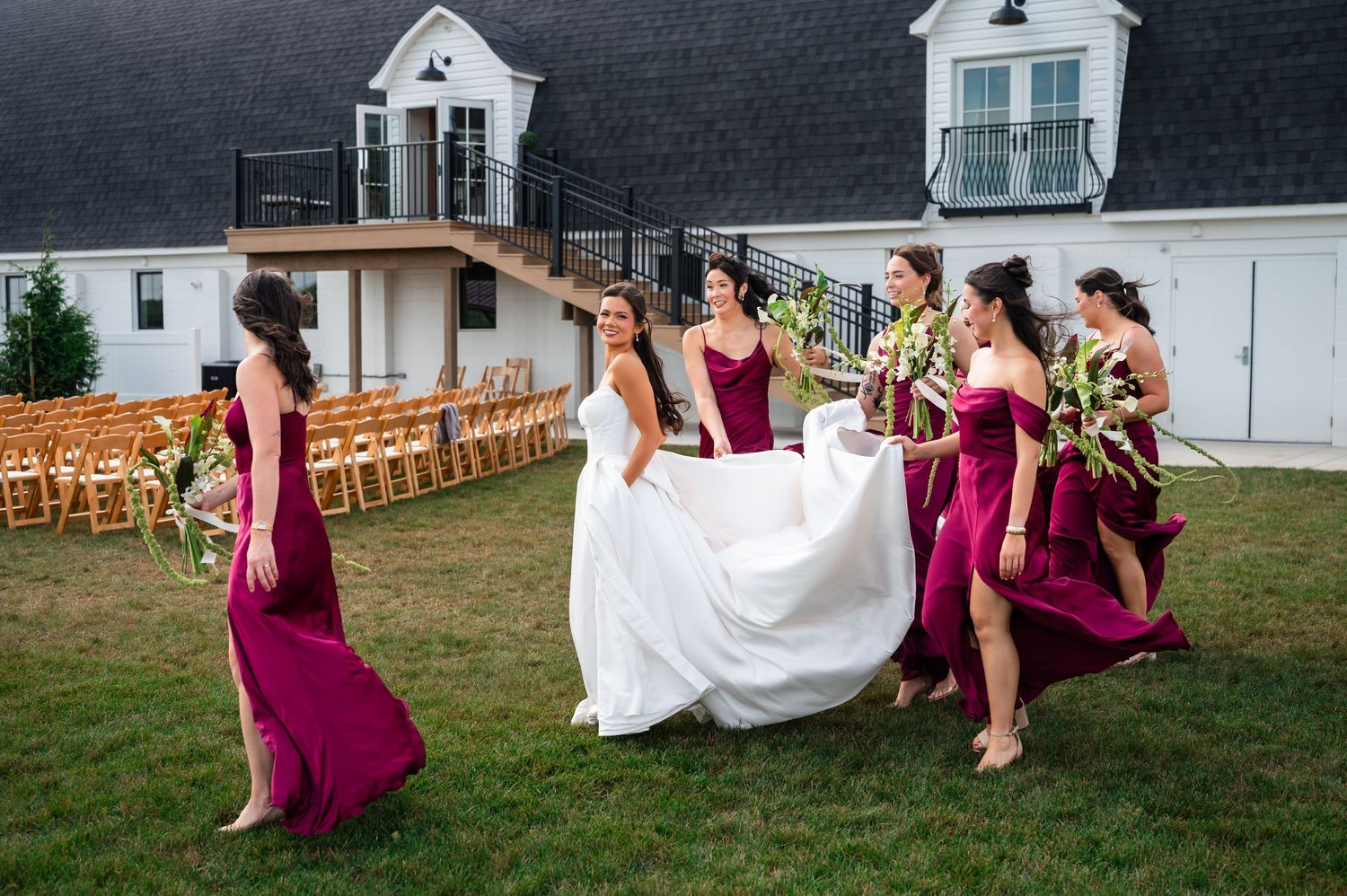 Bride in white gown walks with bridesmaids in burgundy dresses on lawn outside elegant white wedding venue.