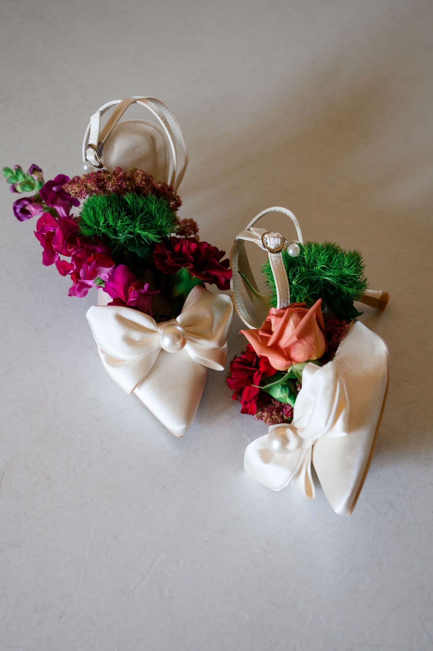 White satin bow bridal heels adorned with colorful fresh flowers including roses and greenery on a neutral background.