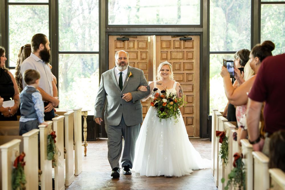 Bride walks down chapel aisle with escort, holding floral bouquet, guests watching in rustic wedding venue.