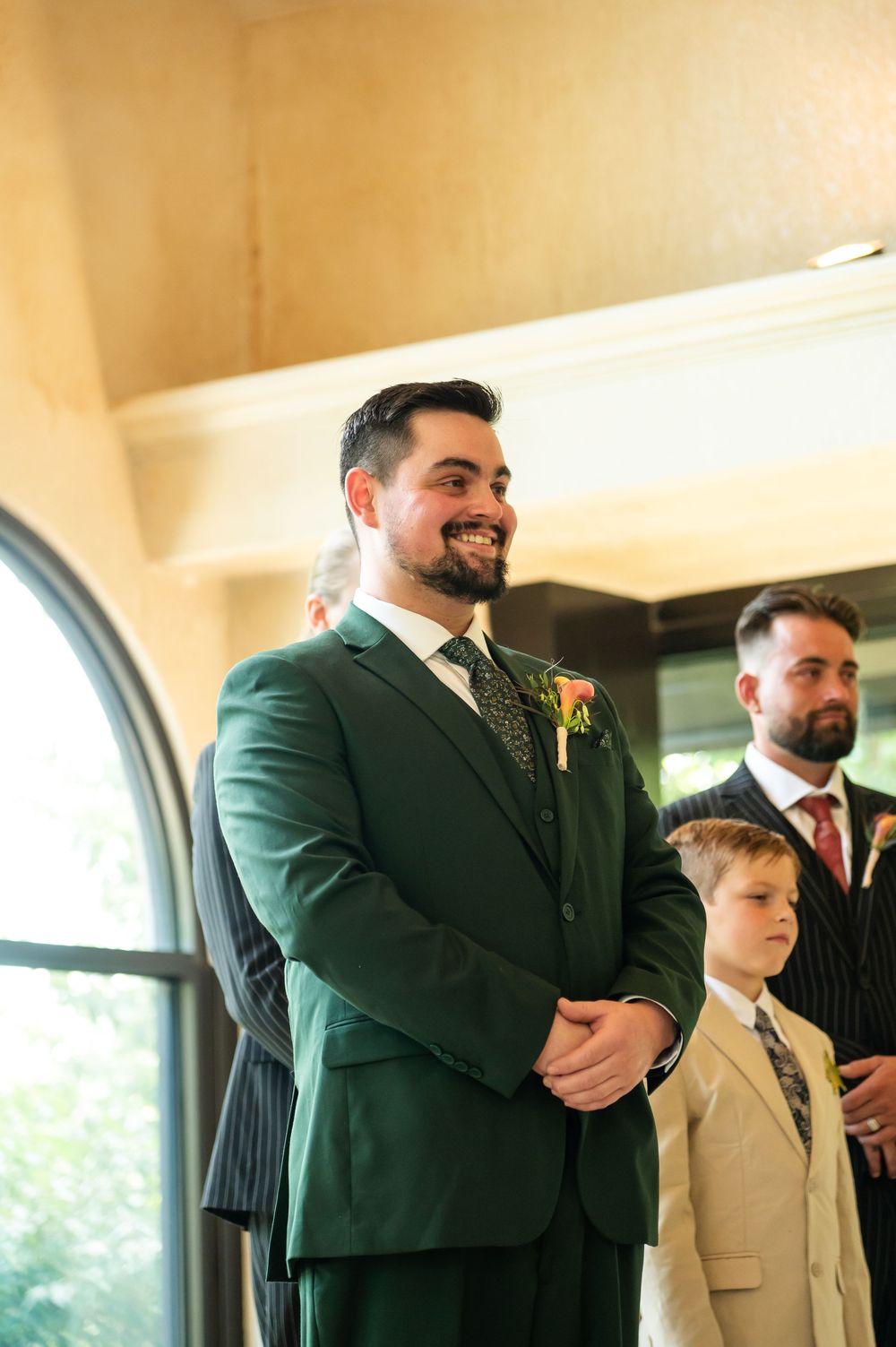Groom in green suit smiling at altar with boutonniere, young boy and groomsman in pinstripe suit beside him.
