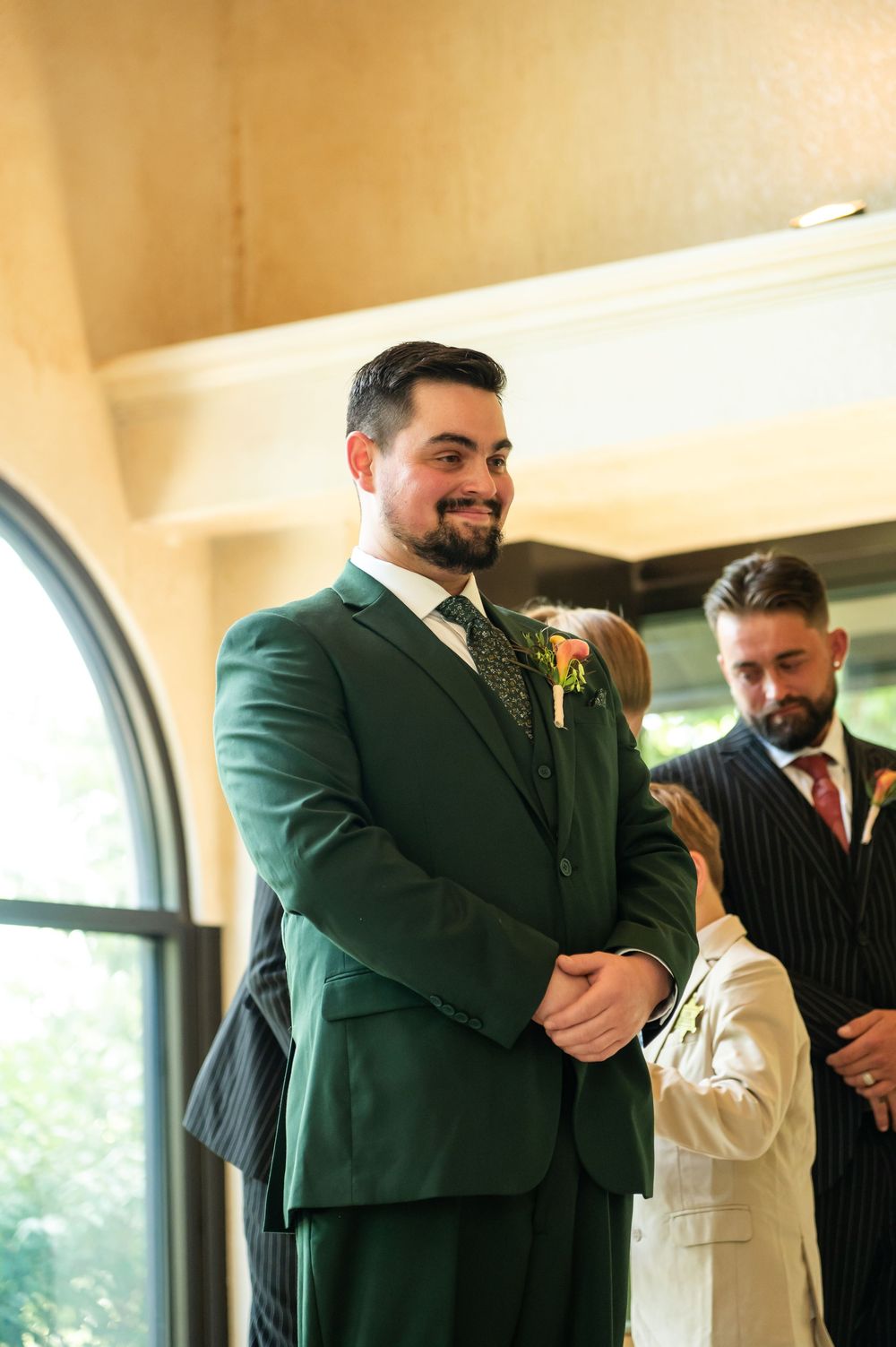 Groom in green suit smiling at wedding ceremony with groomsman in pinstripe suit in background.