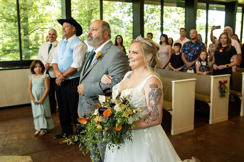 Bride holding colorful bouquet walks down chapel aisle with escort, guests seated in pews during wedding ceremony.