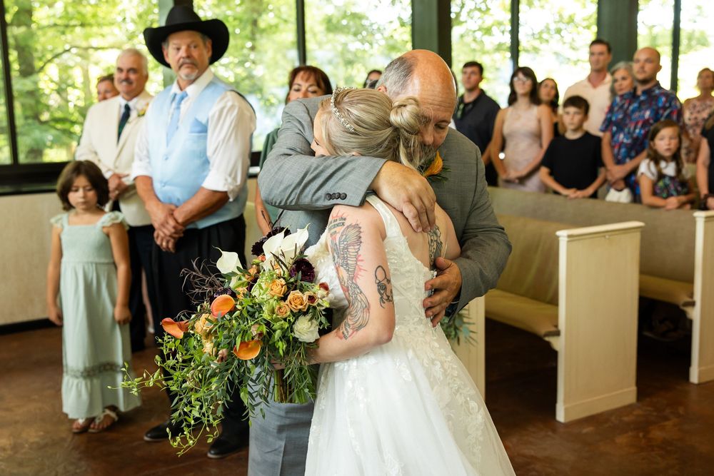 Bride hugging a guest while holding a bouquet of orange and white flowers during a wedding ceremony in a chapel.