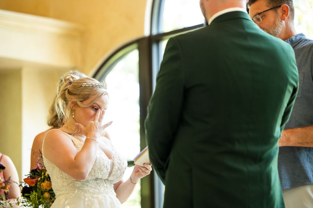 Emotional bride wiping tears during wedding ceremony while groom stands before her in green suit.
