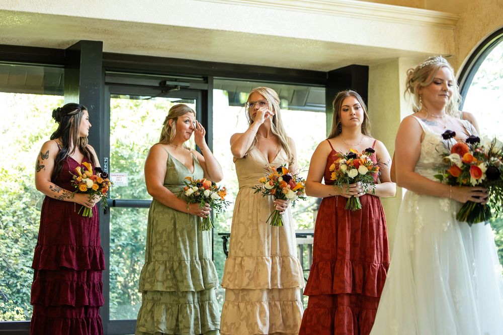 Bridesmaids in mismatched floral dresses hold bouquets as one wipes tears beside a bride in white gown.
