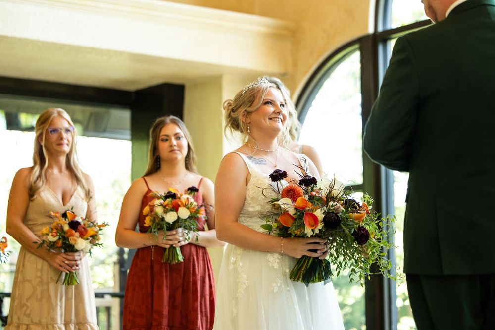Bride holding colorful floral bouquet stands with bridesmaids in mismatched dresses during indoor wedding ceremony.