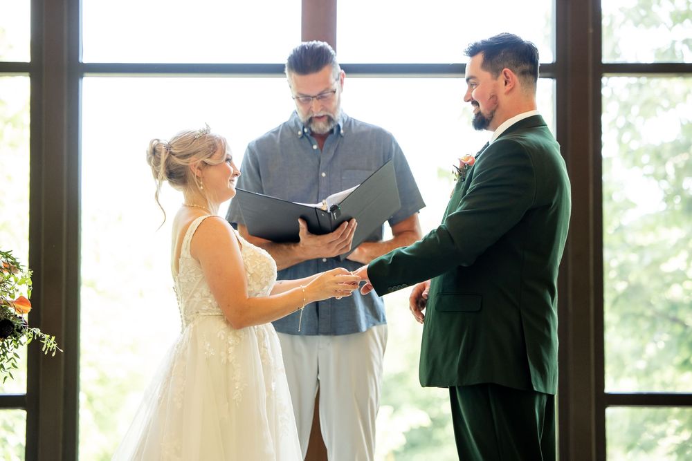 Bride and groom exchange vows with officiant during intimate indoor wedding ceremony near large windows.