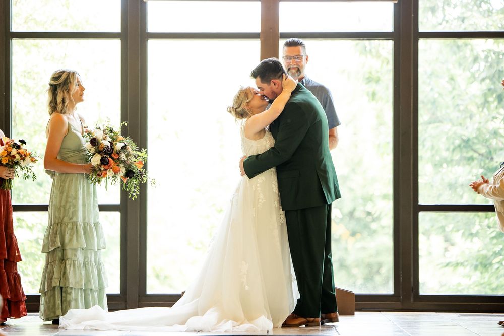 Bride and groom share first kiss at wedding ceremony with officiant and bridesmaid in elegant venue.