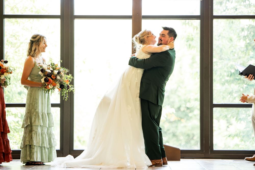 Bride and groom share first dance embrace at wedding ceremony with bridesmaid holding floral bouquet nearby.