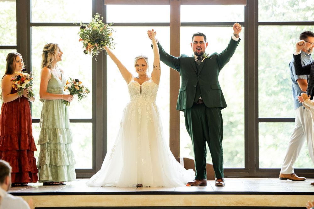 Joyful newlyweds celebrate at wedding altar, bride in white gown raising bouquet, groom in green suit, with wedding party.