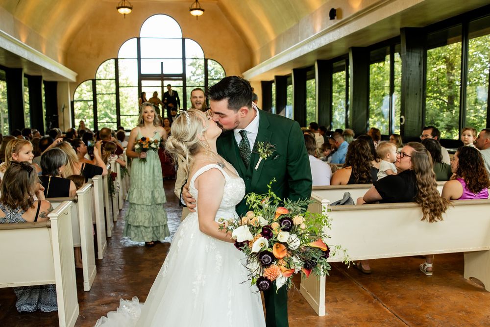 Bride and groom share first kiss in aisle of elegant chapel with guests seated in wooden pews during wedding ceremony.