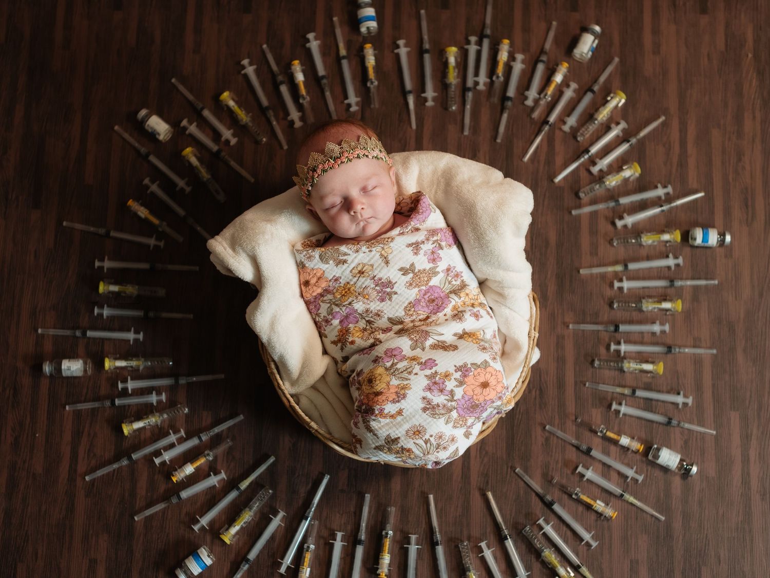 Newborn baby swaddled in floral wrap sleeping in a bowl surrounded by medical syringes on a wooden floor.