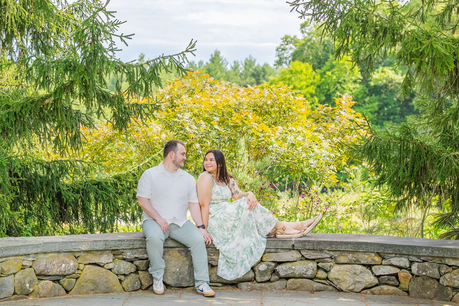 Couple sitting on a stone wall surrounded by lush green trees, smiling at each other in an outdoor garden setting.