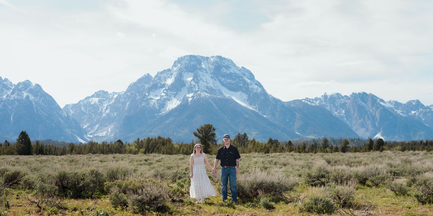 Couple holding hands in a mountain meadow with snow-capped Grand Teton peaks in the background.