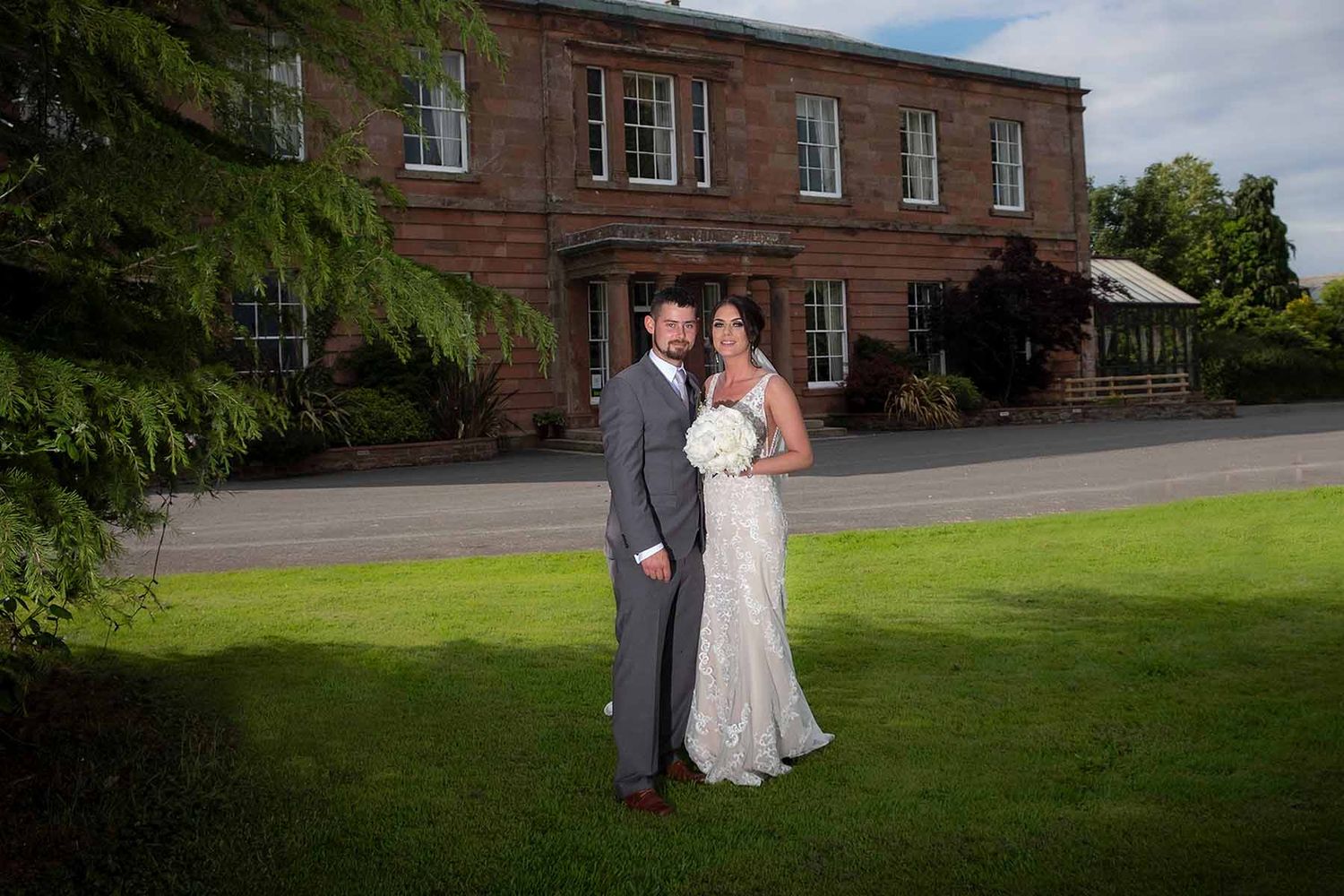 Bride and groom pose together on green lawn in front of Greenhill Hotel wedding venue near Wigton