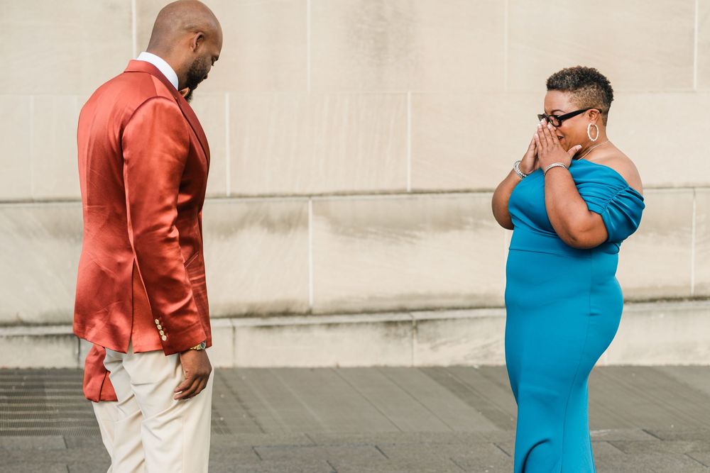 A man in an orange blazer and a woman in a blue dress stand facing each other outdoors.