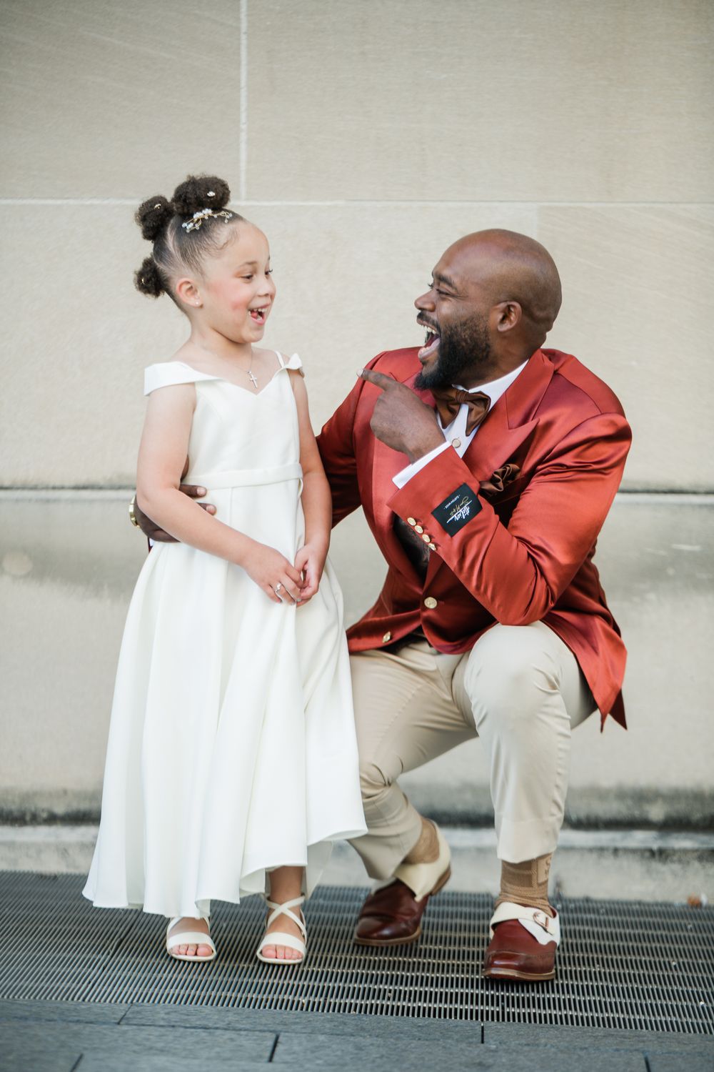 A man in a red blazer crouches to laugh with a young girl in a white flower girl dress at a wedding.