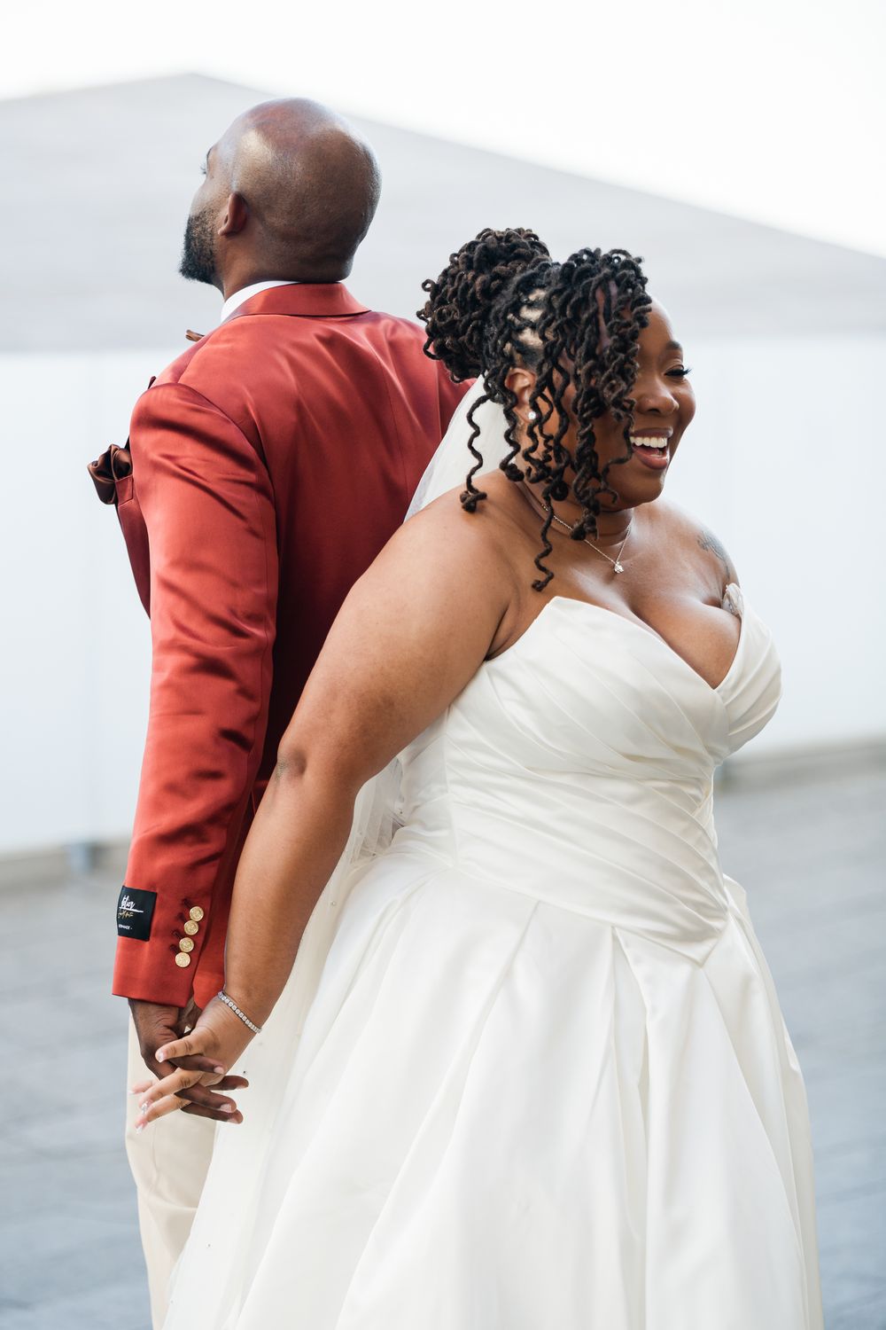 A joyful bride in a white gown holds hands with a groom in a red suit, smiling radiantly during their wedding day.