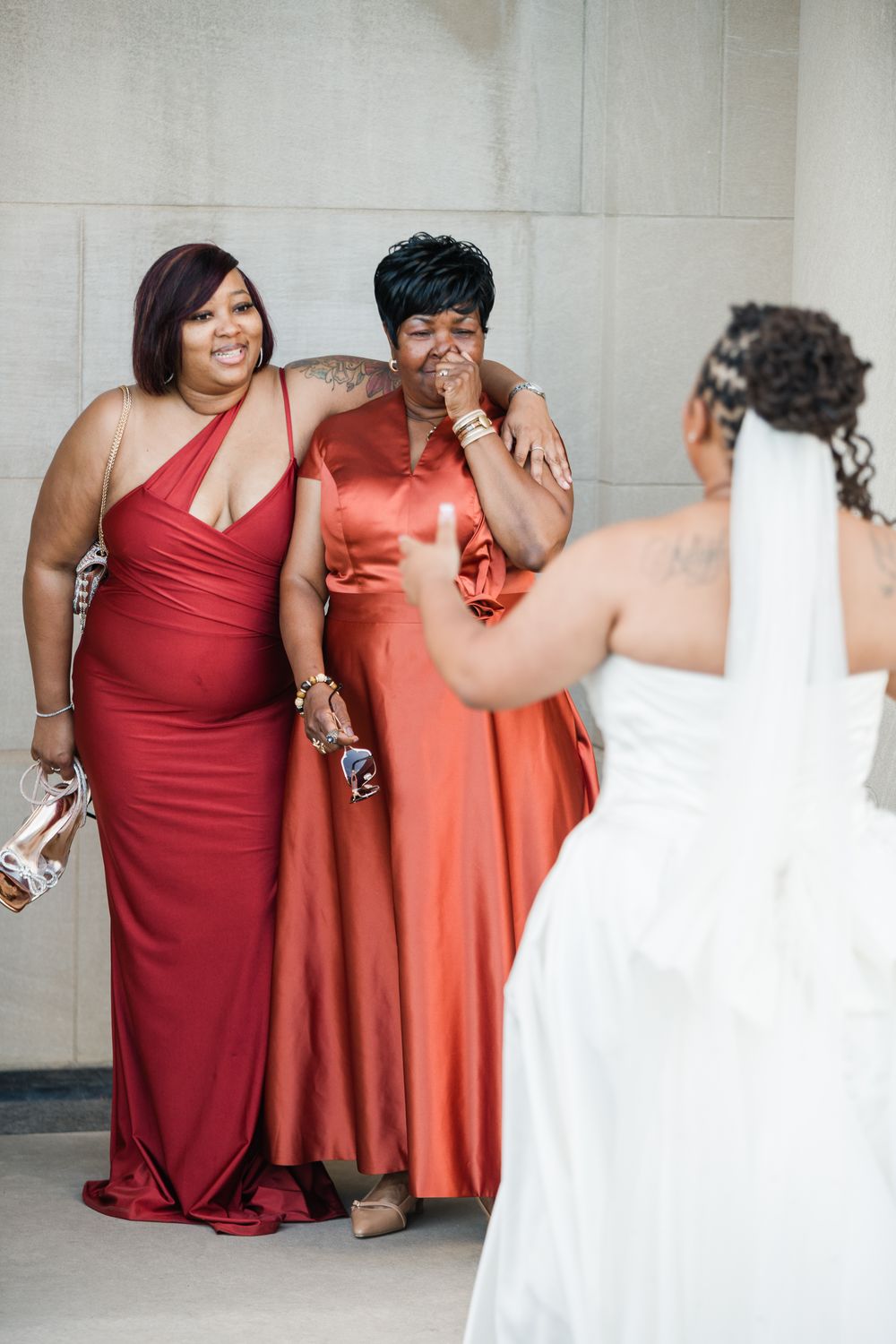 Bride in white gown and veil shares emotional first look moment with two women in red and orange satin bridesmaid dresses.