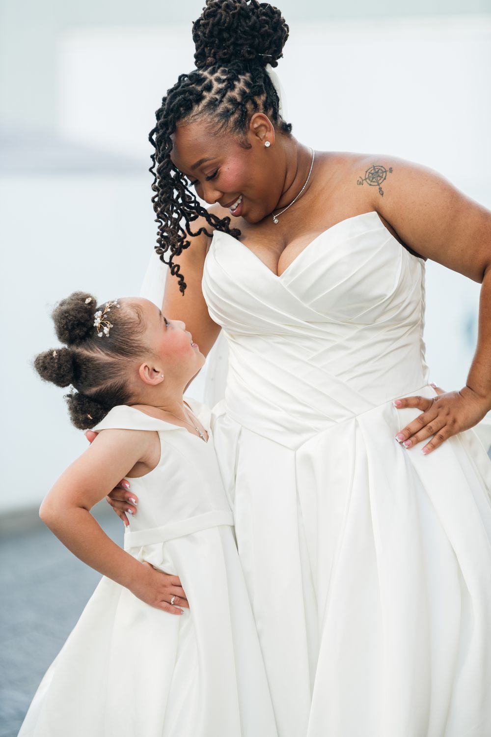 Bride in white gown smiles at flower girl in matching dress, both posing with hands on hips.