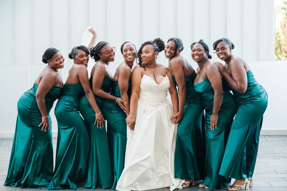Bride in white gown poses with bridesmaids in emerald green satin dresses, laughing together outdoors.