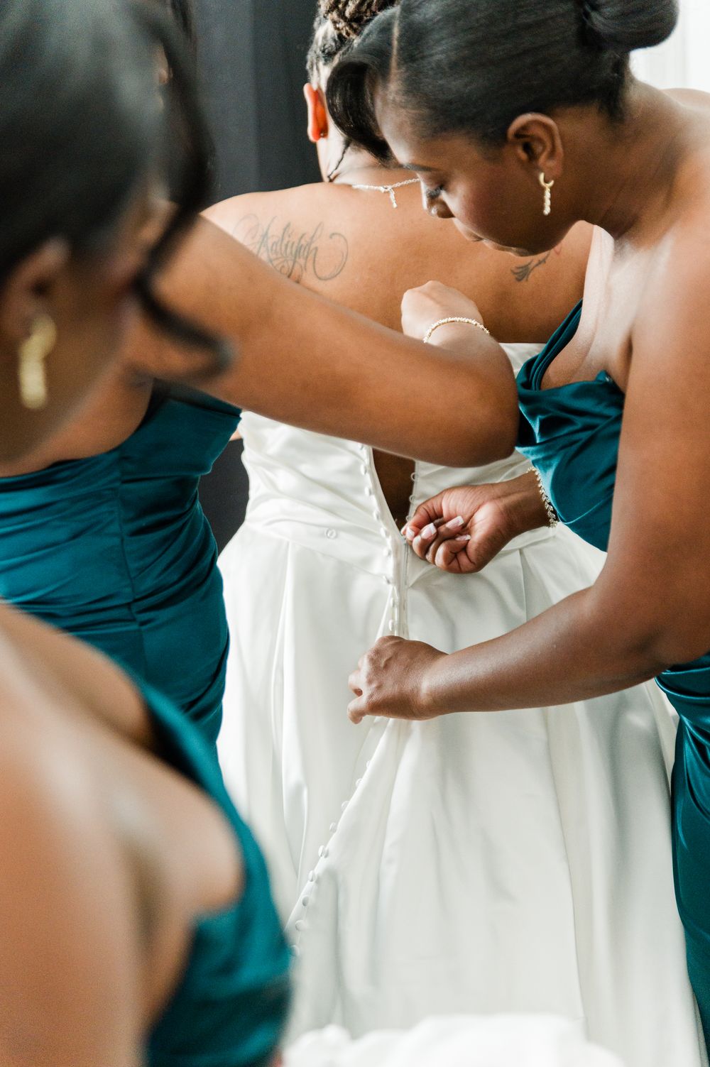 Bridesmaids in teal dresses buttoning the back of a white wedding gown on the bride.