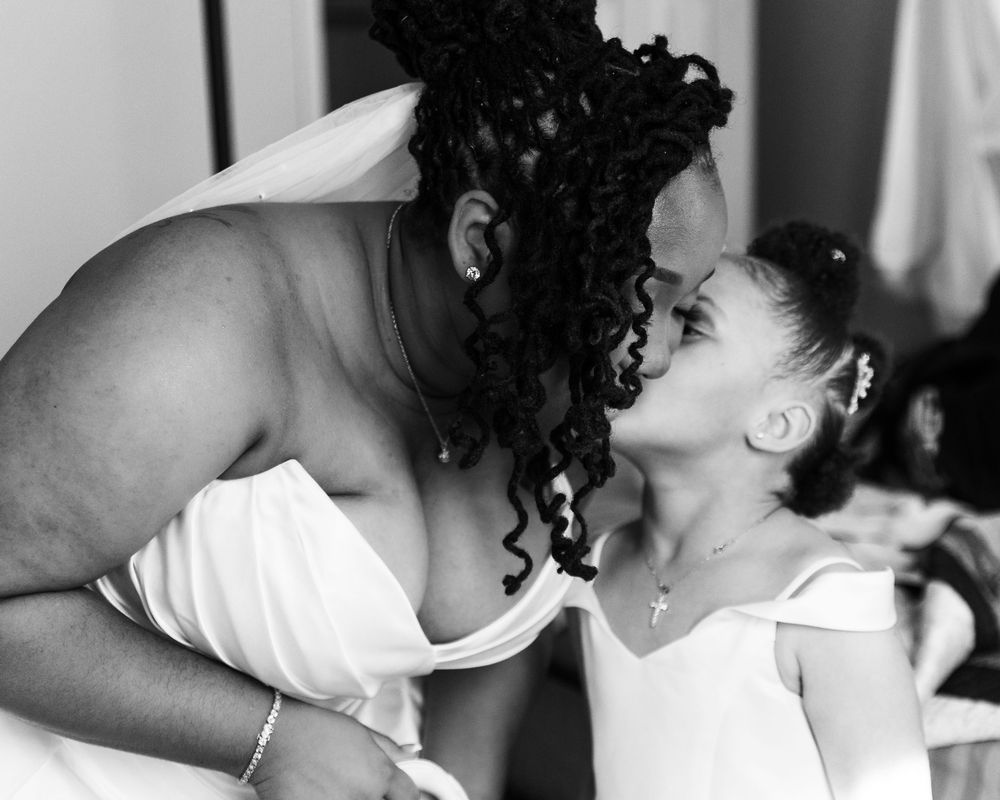 Black and white photo of a bride in a white gown sharing a tender kiss with a young flower girl.