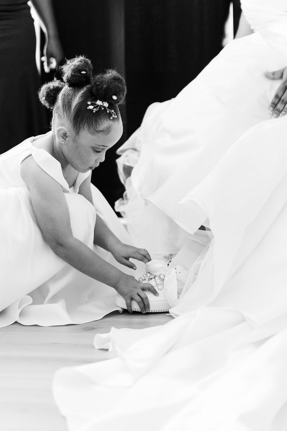 Black and white photo of a young flower girl adjusting a bride's wedding shoe during a ceremony.