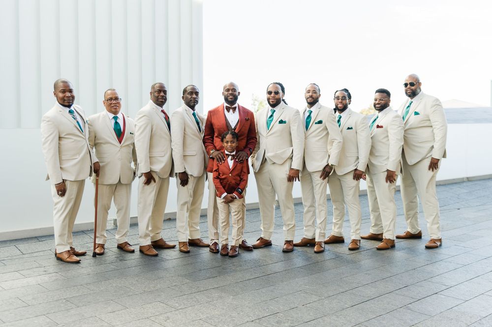 Groom in red suit stands with groomsmen in tan suits and teal ties on rooftop wedding photo.