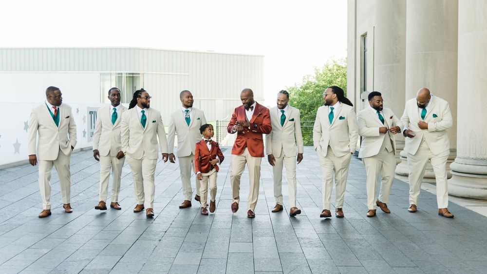 Groom in red jacket poses with groomsmen in cream suits and teal ties outdoors at elegant wedding venue.