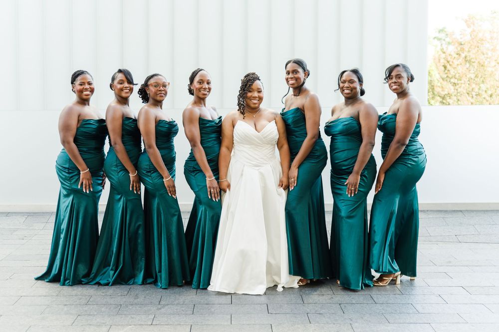 Bride in white gown poses with seven bridesmaids in emerald green satin dresses outdoors.