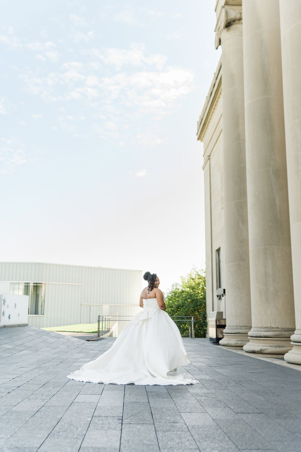 Bride in a flowing white ball gown stands near grand columns of a neoclassical building on a paved plaza.