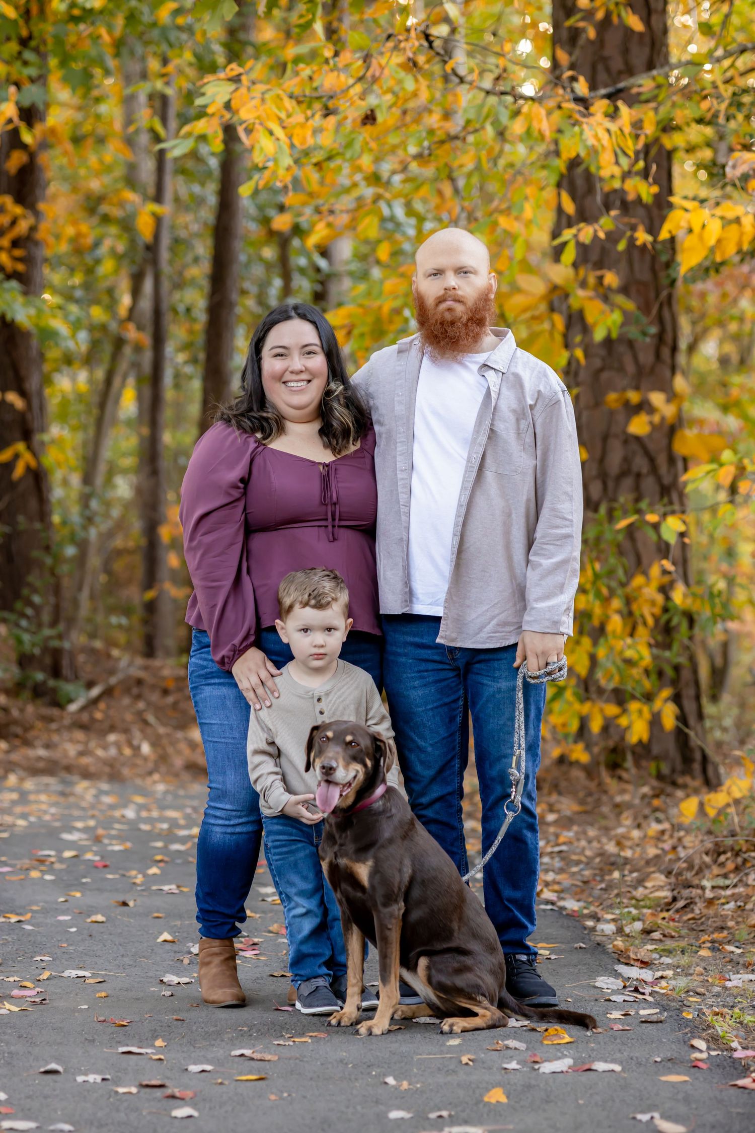 Family of three with red-bearded dad, smiling mom, young boy, and chocolate Lab pose on autumn forest path.