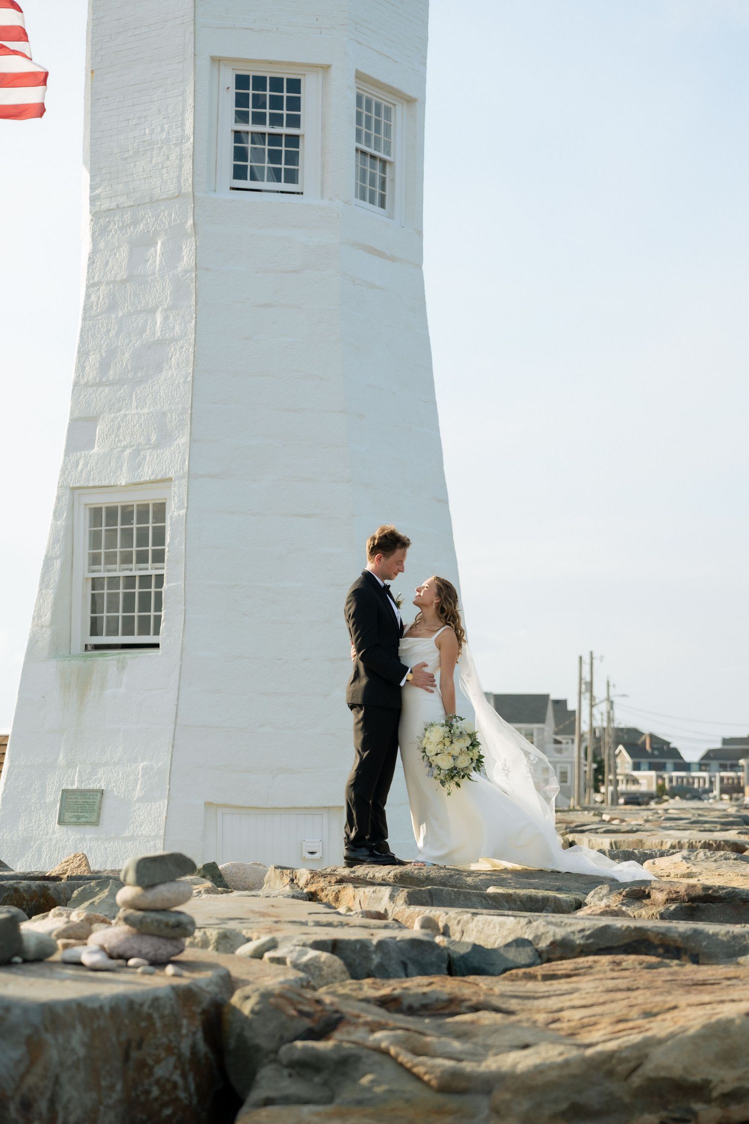 Bride and groom embrace by a white lighthouse on rocky shore, bride holding bouquet, American flag visible.