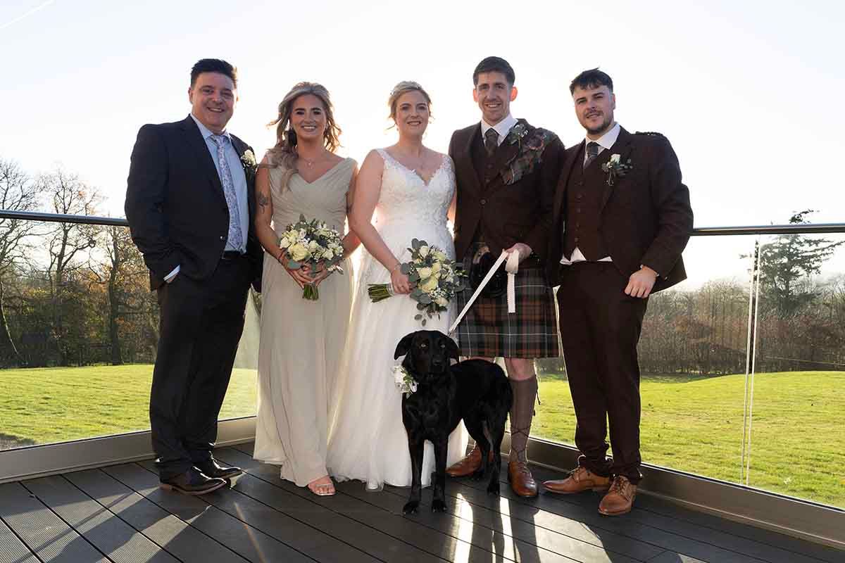 Wedding group photo with Abbie % Ross with three guests, and a black Labrador on an outdoor deck at Hetland Hall