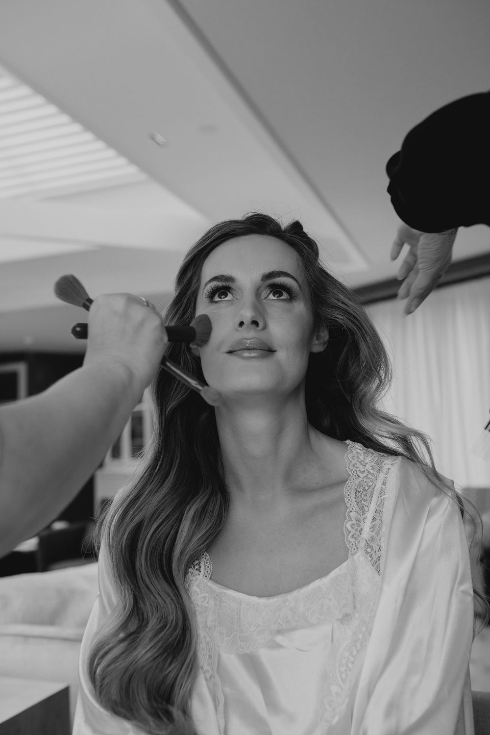 Black and white photo of a bride getting makeup applied with a brush while having her hair styled before her wedding.