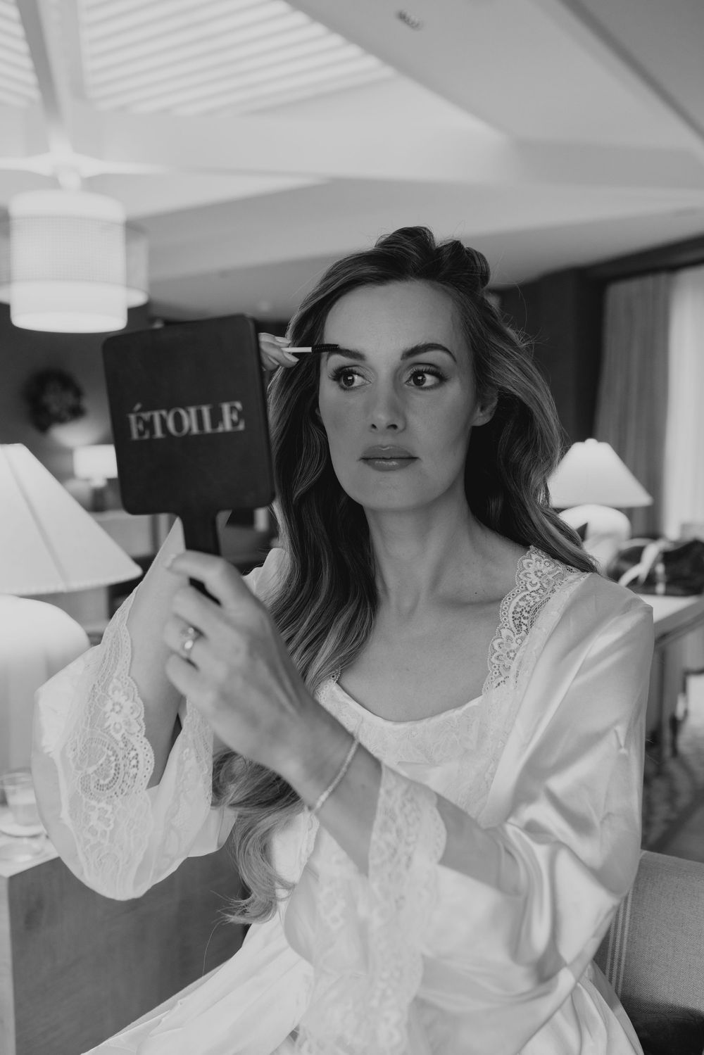 Elegant bride in white lace robe holding an Etoile mirror during wedding preparations, black and white photo.
