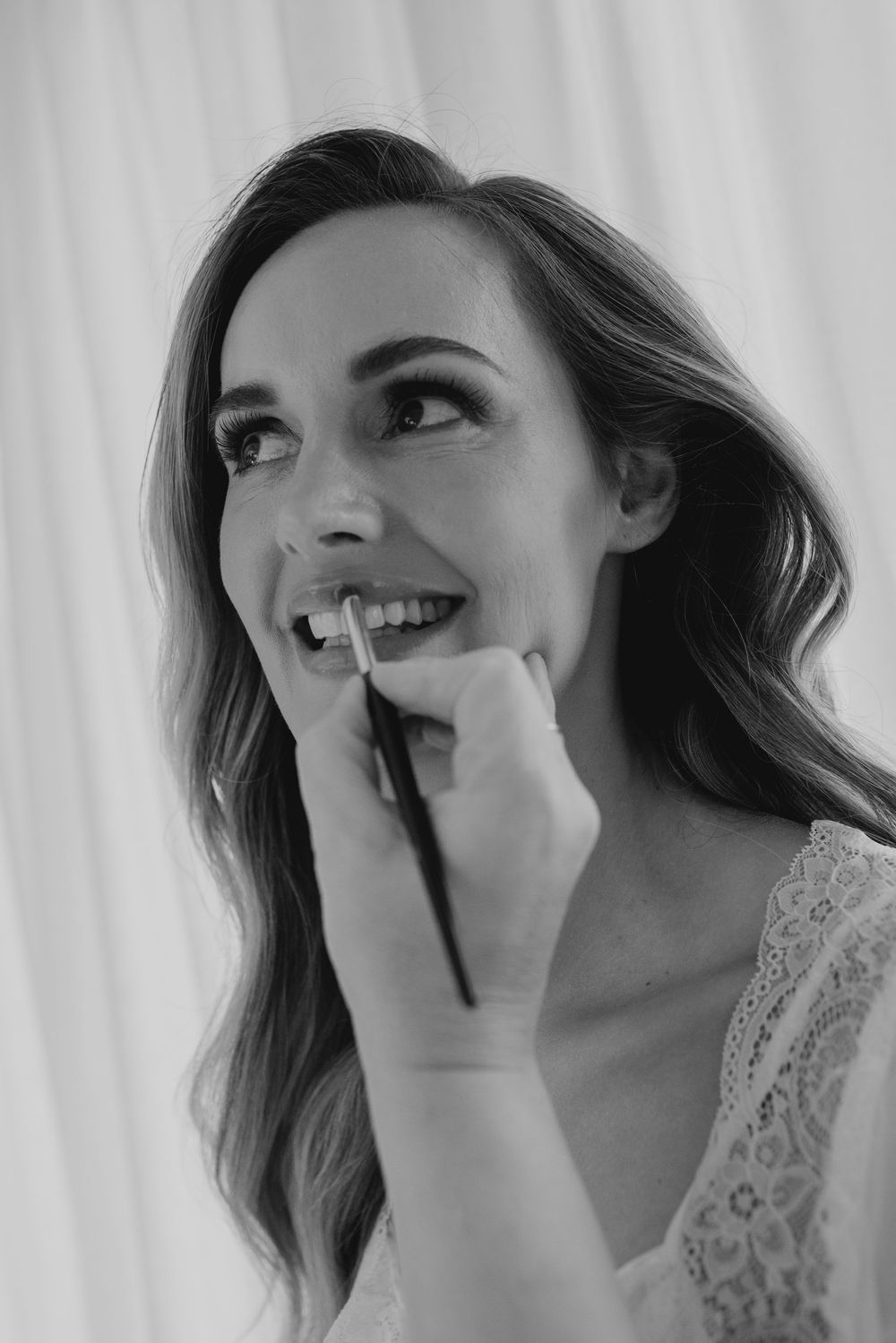 Black and white photo of a smiling bride having lipstick applied by a makeup artist during bridal preparations.