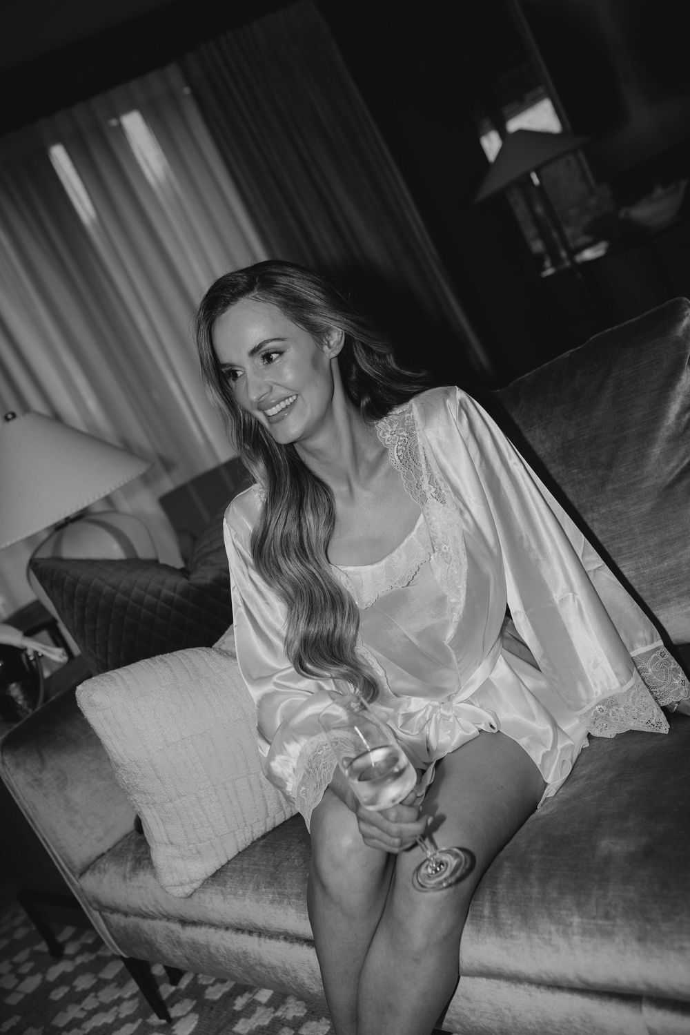 Smiling bride in satin robe holding champagne glass seated on elegant hotel suite chair in black and white photo.